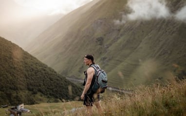 man in gray jacket and black backpack standing on green grass field near mountain during daytime
