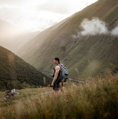 man in gray jacket and black backpack standing on green grass field near mountain during daytime