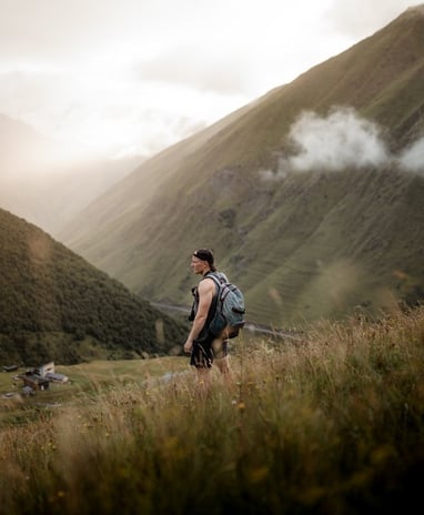 man in gray jacket and black backpack standing on green grass field near mountain during daytime