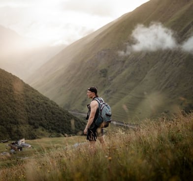 man in gray jacket and black backpack standing on green grass field near mountain during daytime