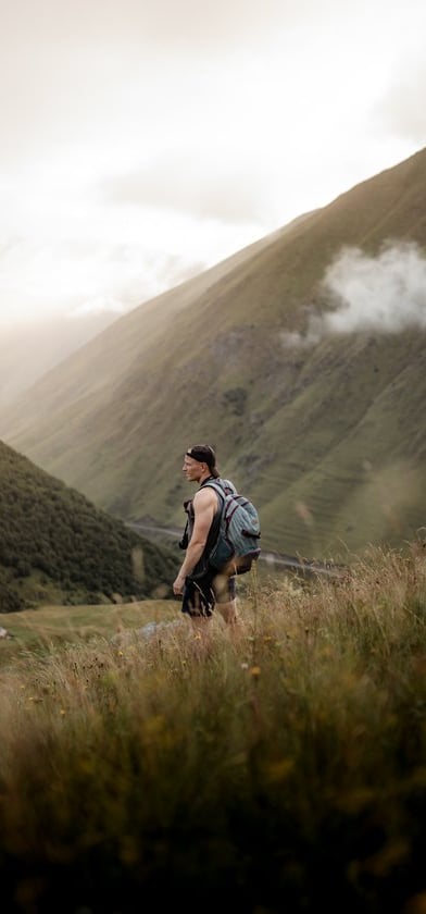 man in gray jacket and black backpack standing on green grass field near mountain during daytime