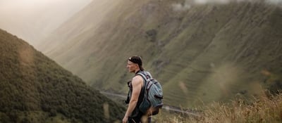 man in gray jacket and black backpack standing on green grass field near mountain during daytime