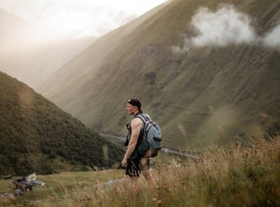 man in gray jacket and black backpack standing on green grass field near mountain during daytime