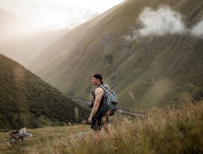 man in gray jacket and black backpack standing on green grass field near mountain during daytime