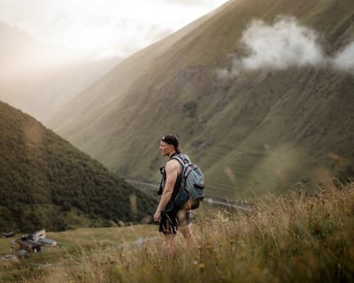 homme en veste grise et sac à dos noir debout sur un champ d'herbe verte près d'une montagne pendant
