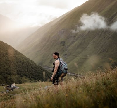 man in gray jacket and black backpack standing on green grass field near mountain during daytime