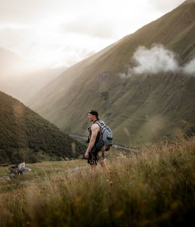 man in gray jacket and black backpack standing on green grass field near mountain during daytime