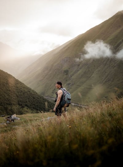 man in gray jacket and black backpack standing on green grass field near mountain during daytime