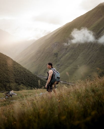 man in gray jacket and black backpack standing on green grass field near mountain during daytime