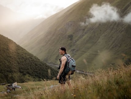 man in gray jacket and black backpack standing on green grass field near mountain during daytime