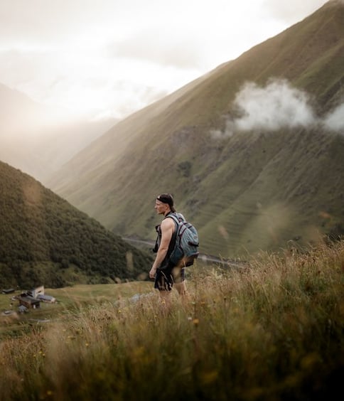 man in gray jacket and black backpack standing on green grass field near mountain during daytime