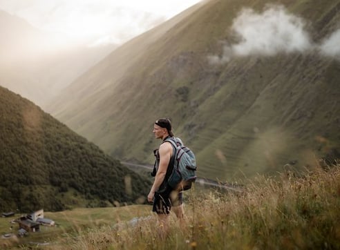 man in gray jacket and black backpack standing on green grass field near mountain during daytime