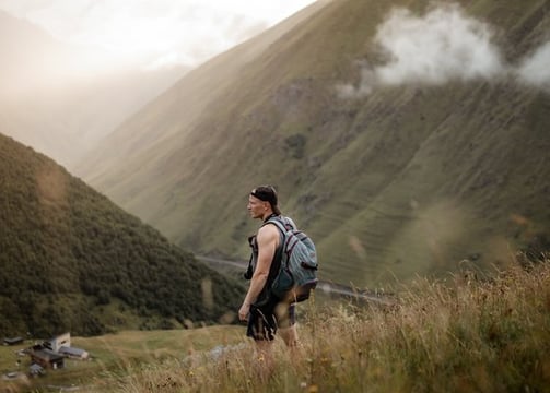 man in gray jacket and black backpack standing on green grass field near mountain during daytime