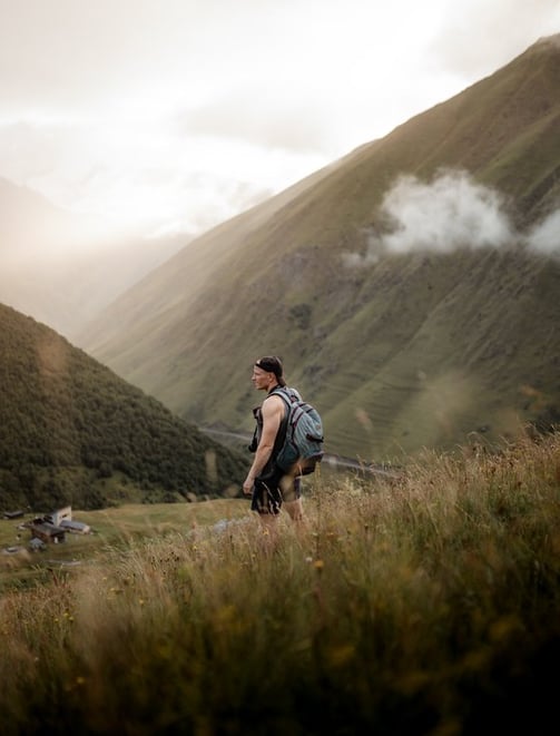 man in gray jacket and black backpack standing on green grass field near mountain during daytime