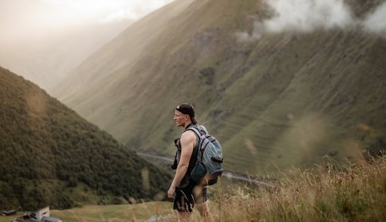 man in gray jacket and black backpack standing on green grass field near mountain during daytime