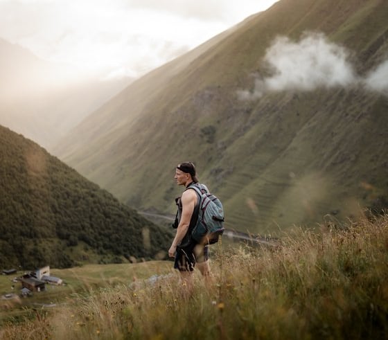 man in gray jacket and black backpack standing on green grass field near mountain during daytime