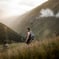 man in gray jacket and black backpack standing on green grass field near mountain during daytime
