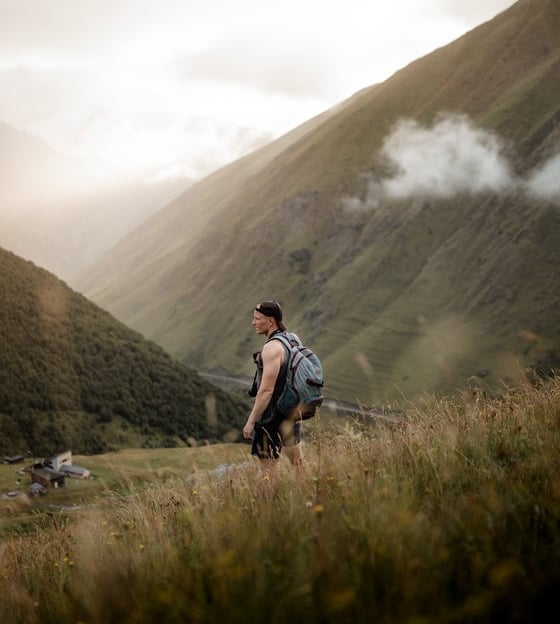 man in gray jacket and black backpack standing on green grass field near mountain during daytime