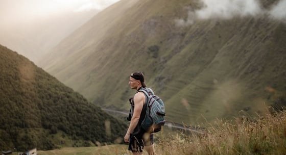 man in gray jacket and black backpack standing on green grass field near mountain during daytime