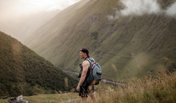 man in gray jacket and black backpack standing on green grass field near mountain during daytime