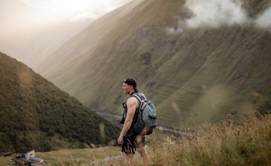 man in gray jacket and black backpack standing on green grass field near mountain during daytime