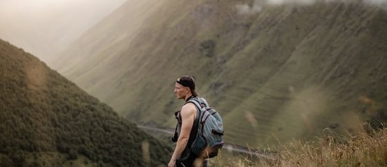 man in gray jacket and black backpack standing on green grass field near mountain during daytime
