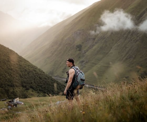 man in gray jacket and black backpack standing on green grass field near mountain during daytime