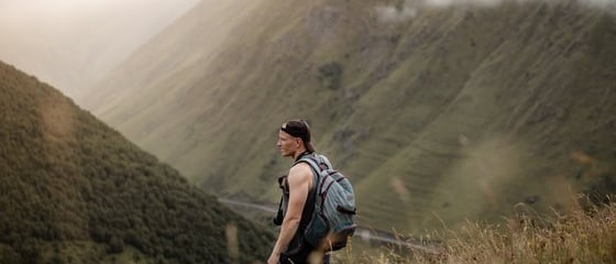 man in gray jacket and black backpack standing on green grass field near mountain during daytime