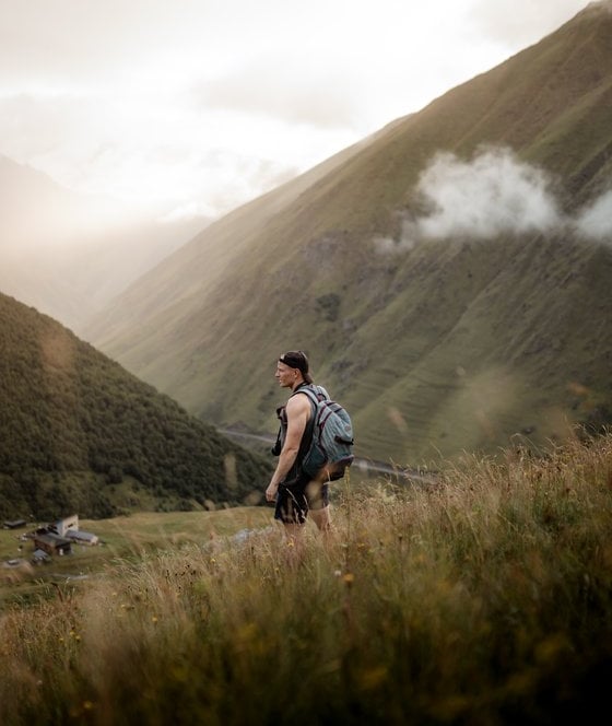man in gray jacket and black backpack standing on green grass field near mountain during daytime