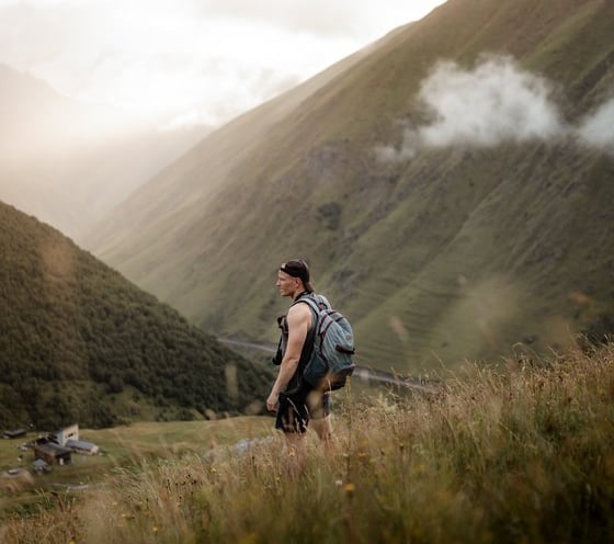 man in gray jacket and black backpack standing on green grass field near mountain during daytime