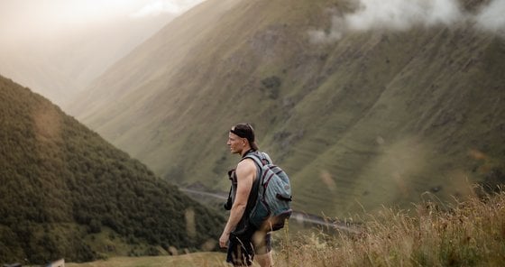 man in gray jacket and black backpack standing on green grass field near mountain during daytime