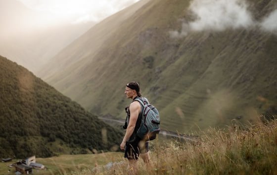 man in gray jacket and black backpack standing on green grass field near mountain during daytime
