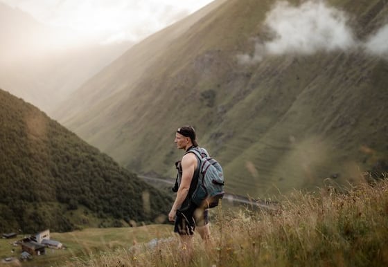 man in gray jacket and black backpack standing on green grass field near mountain during daytime