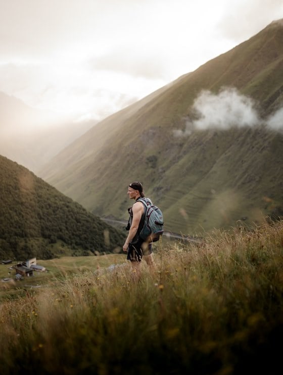man in gray jacket and black backpack standing on green grass field near mountain during daytime