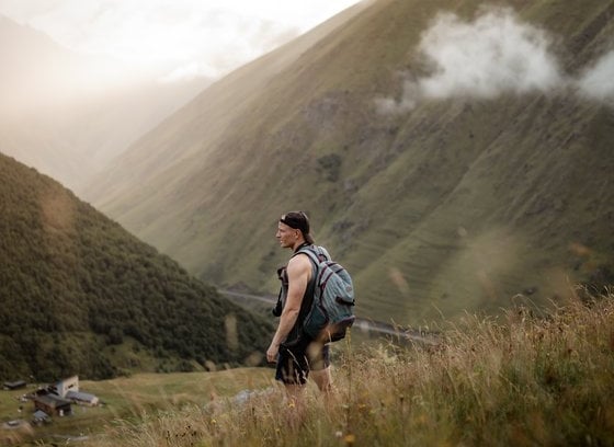man in gray jacket and black backpack standing on green grass field near mountain during daytime