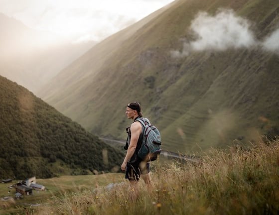man in gray jacket and black backpack standing on green grass field near mountain during daytime