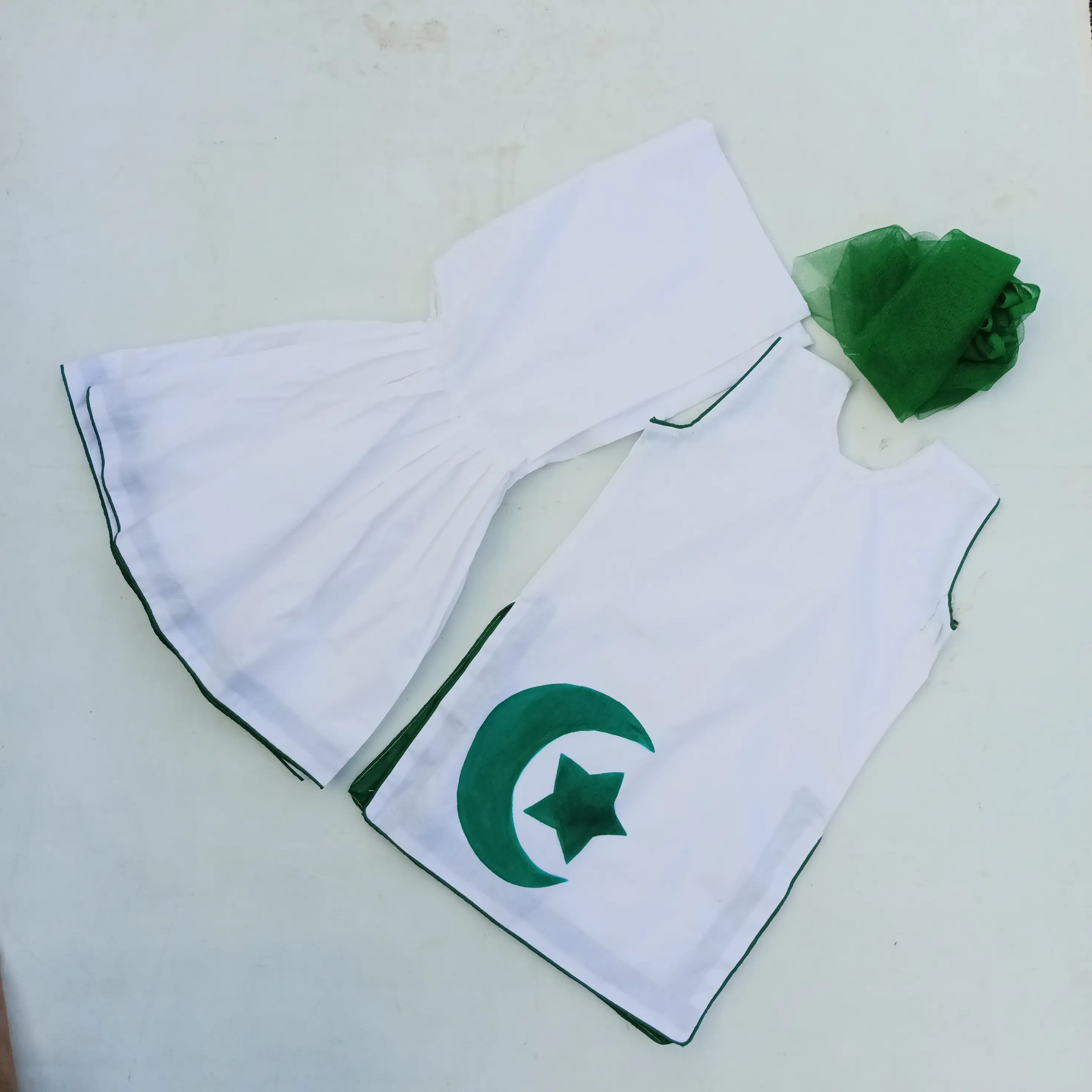 Twirl & Bloom Set A patriotic Pakistani children's dress set features a white cotton sleeveless shirt with a green crescent and star print on the left. It's paired with a matching wide-legged sharara and a sheer green dupatta.