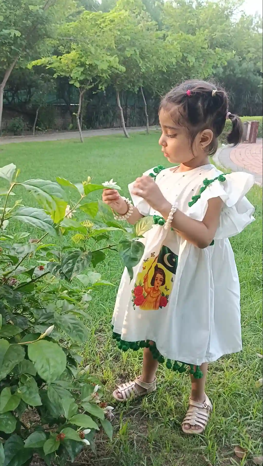 Starry Blossom Frock Patriotic Elegance for Little Hearts "A young girl in a white Pakistani Independence Day dress, holding a white flower in a garden."